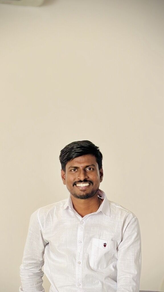 Smiling person in a white button-down shirt, seated against a plain cream wall